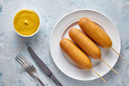 Corn dog traditional American street junk food deep fried hotdog meat sausage with mustard snack on stick coated in a thick layer of cornmeal batter unhealthy eating on rustic table.の写真素材