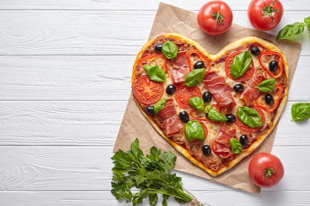 Heart shaped pizza love concept Valentines Day symbol romantic dinner Italian food. Prosciutto, olives, tomatoes, parsley, basil and mozzarella cheese meal served on white wooden table backgroundの写真素材