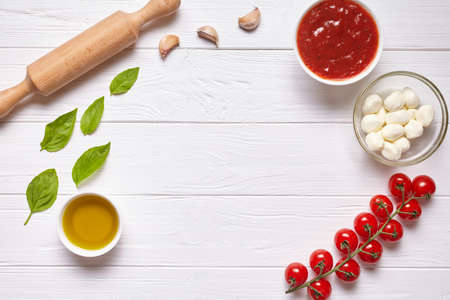 Rustic traditional Italian food background with empty copy design space on white wooden texture kitchen table. Basil, olive oil, tomato sauce, rolling pin, tomatoes. Flat lay, top view.の写真素材