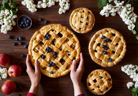Homemade organic apple pie pies ready to eat hold female hands on dark wooden table with flowers, blueberry and apples. Traditional dessert on Independence Day. Food flat lay backgroundの写真素材