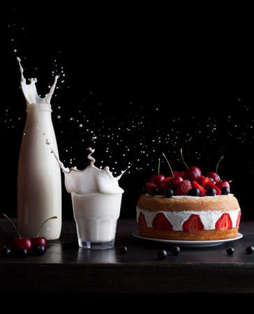 Splashes of milk in a glass and jar and biscuit sponge cake with strawberries, cherry and currrant on a black background. Frozen motion. Creative food photo. Summer delicious dessert.の写真素材