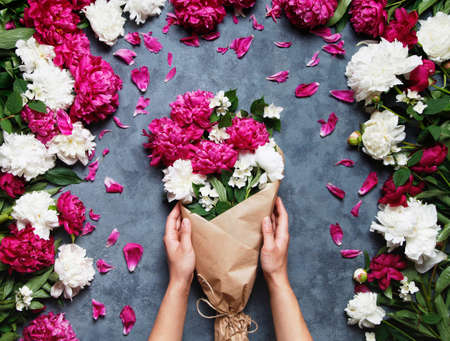 Top view of hands of young woman florist holding beautiful bouquet of peonies. Florist at work: pretty woman making summer bouquet of peonies on a working gray table. Flat lay composition.の写真素材