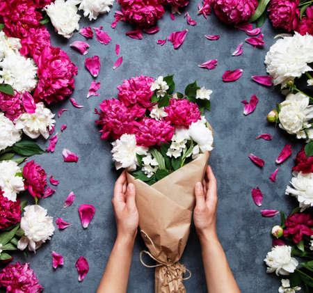 Top view of hands of young woman holding beautiful bouquet of peonies. Florist at work: pretty woman making summer bouquet of peonies on a working gray table. Flat lay composition.の写真素材