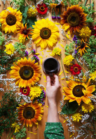 Floral summer background. A mug of coffee in a womans hand on a wooden background with sunflowers and wildflowers flowers. Hello summer July, August. Arrival of summer mood. Flat lay.の写真素材