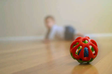 Plastic ball toy on the wooden floor with blurred crawling baby in backgroundの素材