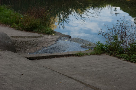 water flows out of a pipe in a concrete dam, the grass is green all around, and the blue sky is reflected in the waterの写真素材