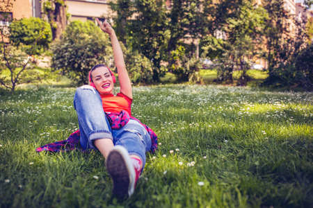 Smiling urban female with earphones and smartphone listening to music on grass in parkの写真素材