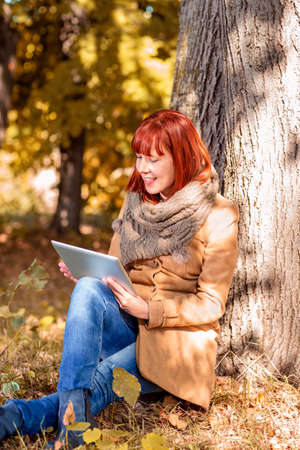 Young woman sitting under a tree in autumn park with tabletの写真素材