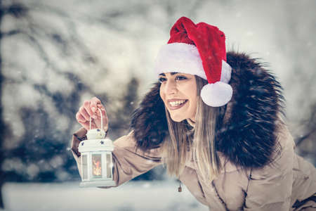 Young happy woman in Santa hat holding Christmas lantern outdoors on beautiful winter snow day - Christmas timeの写真素材