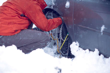 Young Man preparing car for travelling at winter day. Snow chains on the wheels of car snowy day.