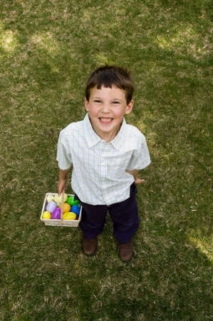 a boy stands on the lawn holding his white easter basket full of colorful plastic eggs after an...