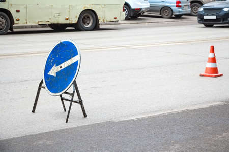 Close up round blue road sign with arrow to the left meaning detour because of road works and an orange cone in backgroundの写真素材