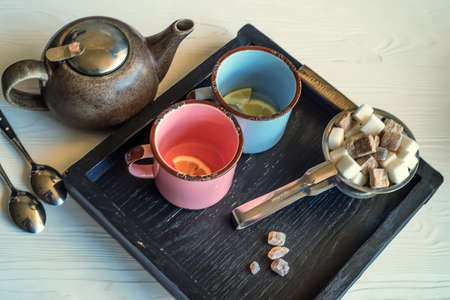 Close up vintage tea set with blue and pink metal mugs, kettle, sugar-tongs and lumps of sugar on wooden trayの写真素材