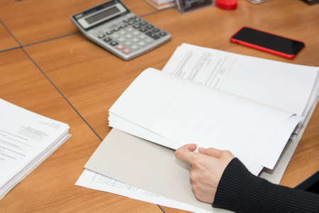 Close-up image of a desk with calculator, mobile phone and documents on it and arms of business woman reading themの写真素材