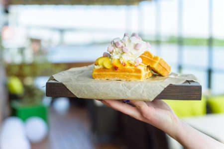 Closeup of waiter's hand holding fresh belgian waffles with mango and cream on wooden tray in...