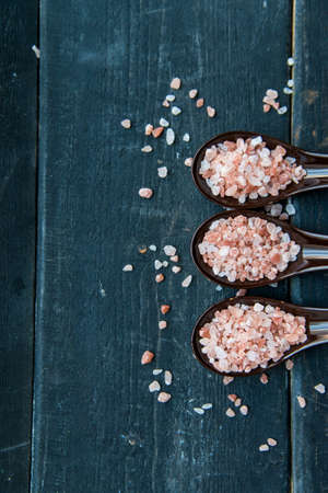 Top view of three spoons with rough pink salt on dark wooden background