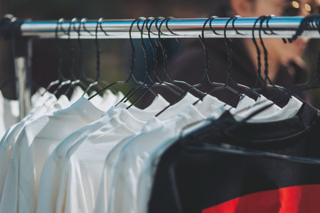 White t-shirts and a single black shirt with a red element are displayed on black hangers from a metal rack. The scene suggests an outdoor market or clothing sale in the daylight.