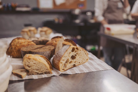 Slices of sourdough bread rest on a kitchen counter while bakery staff work on preparing more dough and baking other varieties of bread nearby.
