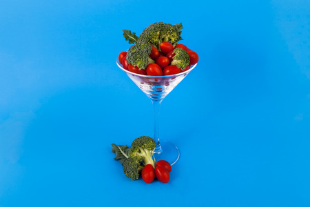 Fancy martini beverage glass filled with sweet cherry tomatoes broccoli florets on a solid blue backgroundの写真素材