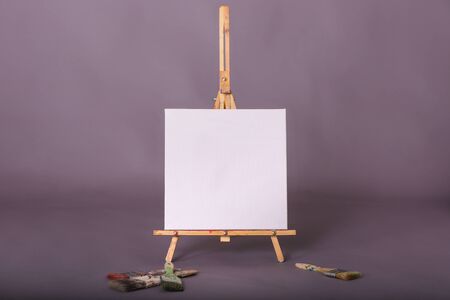 Blank square white painters canvas sitting on artist easel paintbrushes scattered in studioの写真素材