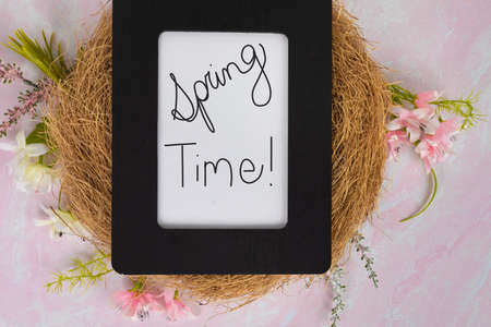 Spring time! Sign in black picture frame laying on a bird nest with pink and white flowers flat lay backgroundの写真素材