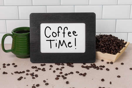 Coffee Time! Sign handwritten on white paper sign in black picture frame sitting by scattered and piled up whole roasted coffee beans and a green ceramic cupの写真素材