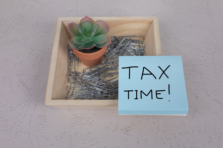 Tax Time! Handwritten sign one light blue square posted notepad with small office desk plant and paperclips accountant workspaceの写真素材