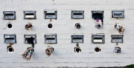 Two rows of old rustic mailboxes in a brick wall painted white with newspapers sticking out.の写真素材