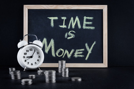 Vintage alarm clock showing 5 o'clock and coin towers with chalckboard and "time is money" text on black background.の写真素材