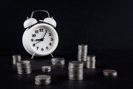 Vintage alarm clock showing 9 o'clock and coin towers on black background. Time is money business conceptの写真素材