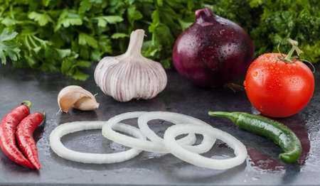 Hot chilli peppers, garlic, purple onion, truss tomato and parsley. Wet raw food ingredients on stone background. Shallow depth of field.の写真素材