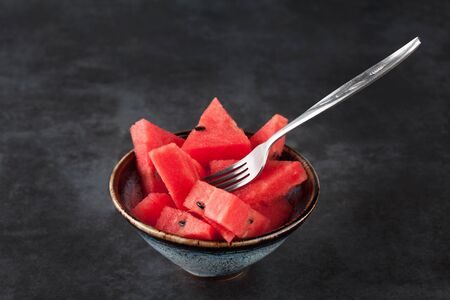 Sliced watermelon in ceramic bowl with fork on dark grungy background with copy space. Shallow depth of field.の写真素材