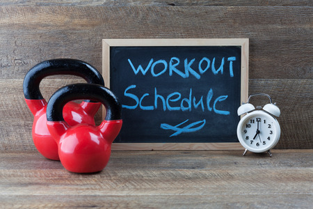 Two red kettlebells with vintage clock and small blackboard on wooden table. Workout schedule concept.の写真素材