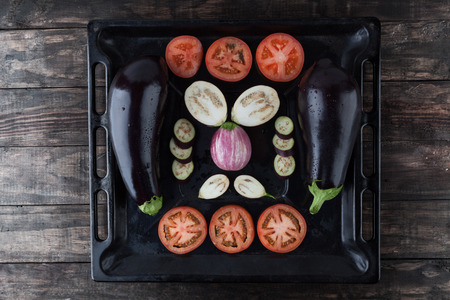 Whole and sliced eggplants and tomatoes on rustic baking tray. Top view.の写真素材