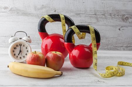 Two red kettlebells with measuring tape, apples, banana, and vintage clock on rustic white wooden table. Healthy diet and fitness concept.の写真素材