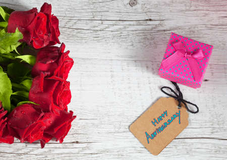 Happy Anniversary text with small gift box and red roses on white rustic wooden table.の写真素材