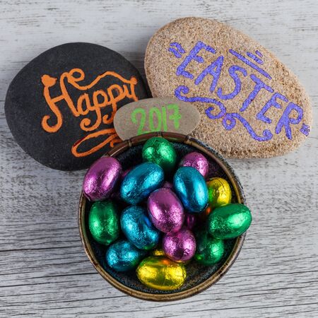 Happy Easter 2017 lettering written on pebbles with chocolate eggs wrapped in colorful foil. Square...