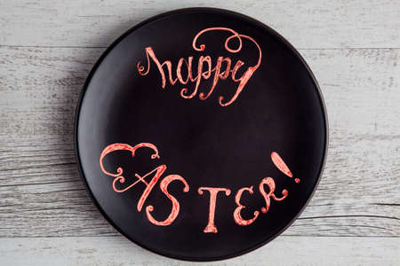 Happy Easter phrase hand written on black plate placed on white rustic wooden table with copy spaceの写真素材
