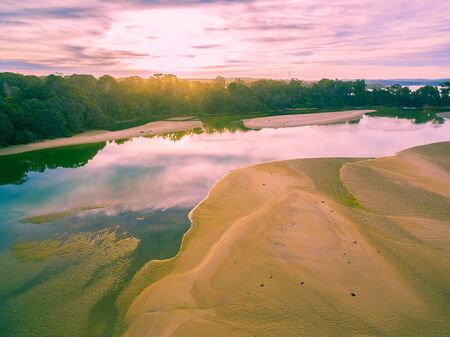 Aerial view of shallow ocean water, sand, and lush coastline vegetation at sunst