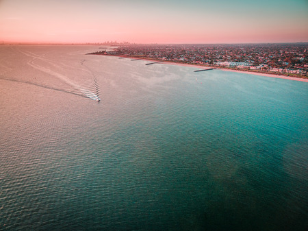 Aerial view of boat sailing across Port Phillip bay with Melbourne coastline and suburban areas in the background at beautiful sunsetの写真素材