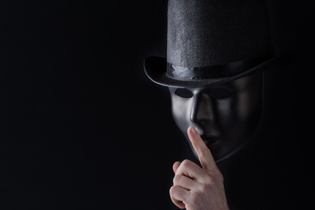 Male finger showing shh sign on black mask wearing black top hat on black background with copy space. Freedom of speech and silence conceptの写真素材
