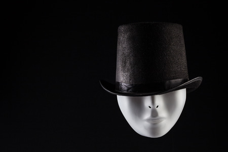 White maske hiding eyes under black top hat isolated on black background with copy space. Hidden personality conceptの写真素材