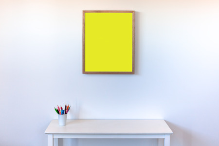 Empty picture frame hanging above white side table with colorful pencils in cup against white wall with copy spaceの写真素材