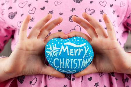 Young girl forming heart shape with fingers holding painted rock - Merry Christmas greetingの写真素材