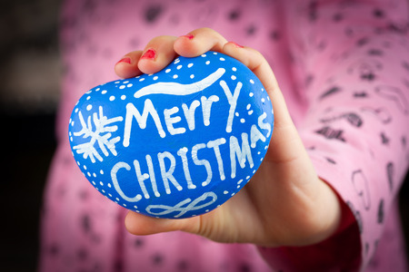 Young girl hand showing blue painted stone with Merry Christmas hand lettering on blurred backgroundの写真素材