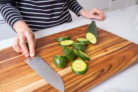 Caucasian woman holding chef knife on wooden cutting board with sliced cucumber in modern kitchen interiorの写真素材