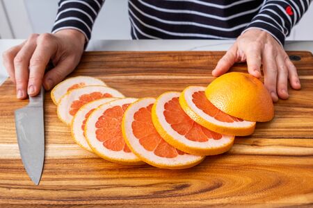 Caucasian woman hands placed on wooden chopping board with sliced red grapefruit and knifeの写真素材
