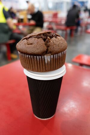 Chocolate muffin placed on top of brown coffee cup on red table with blurred people in the backgroundの写真素材