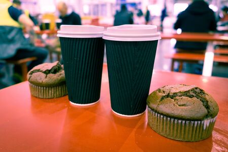 Two muffins and tall coffee cups on a table with blurred people in the backgroundの写真素材