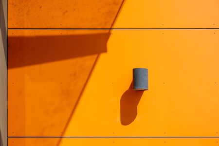 Exterior industrial wall mounted cylinder lamp on modern orange wall with strong shadows in bright daylight and copy spaceの写真素材